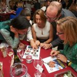 Four adults, gathered around a table full of drinks, quiz cards and part poppers, have their heads together as they try to answer a quiz question.