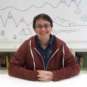 Linden is a white person with short, straight brown hair tucked behind their ears. They are smiling broadly at the camera with their hands interlaced on the table in front of them. They are wearing glasses, a blue shirt and a maroon hoodie.