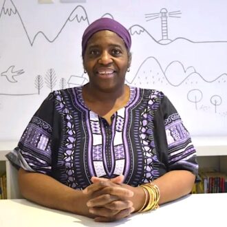 Sandra Agard is a black woman, she sits smiling with her hands intertwined on a white desk. She is wearing a deep purple and white patterned top with a purple head wrap.
