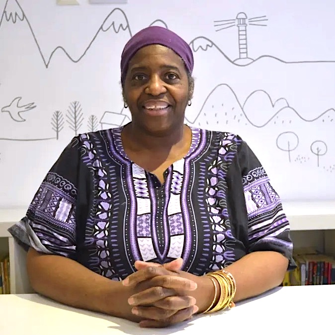 Sandra Agard is a black woman, she sits smiling with her hands intertwined on a white desk. She is wearing a deep purple and white patterned top with a purple head wrap.