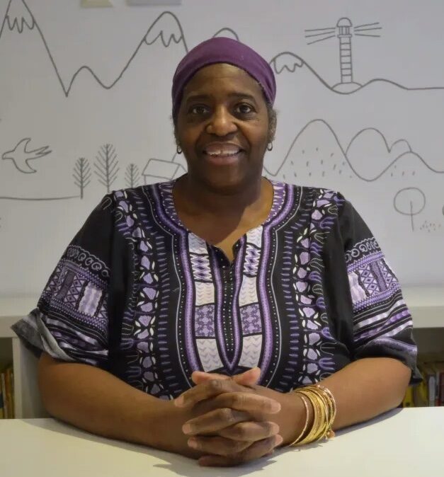 Sandra sits smiling with her hands intertwined on a white desk. She is wearing a deep purple and white patterned top with a purple head wrap.