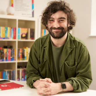 Oliver - a white man with dark, curly hair and thick beard - sits with his hands clasped in front of him. He wears a green t-shirt and green cord jacket and smiles cheerfully at the camera.