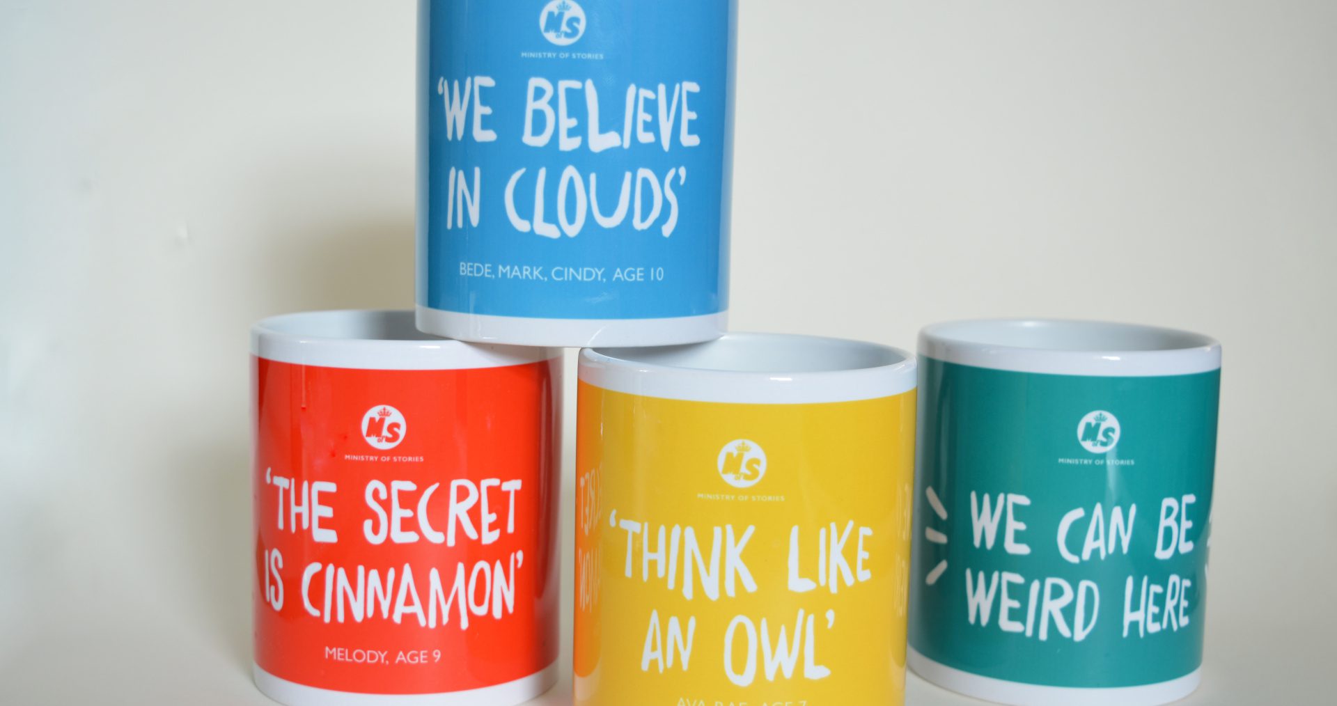 Four mugs in bright colours, featuring the four quotes "we can be weird here", "we believe in clouds", "the secret is cinnamon" and "think like an owl"