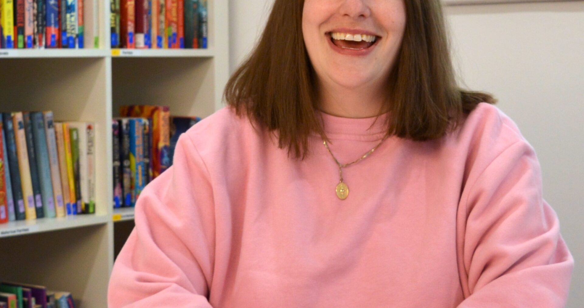 Thea is a white woman with shoulder length dark hair, she's sitting behind a desk wearing a light pink jumper and a short necklace. She is smiling brightly at the camera and has her hands folded on the desk in front of her.