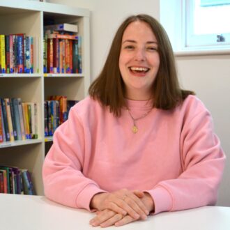 Thea is a white woman with shoulder length dark hair, she's sitting behind a desk wearing a light pink jumper and a short necklace. She is smiling brightly at the camera and has her hands folded on the desk in front of her.