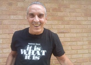 Joe smiles at the camera standing in front of a brick wall, wearing a Parkinson's Disease fundraising t-shirt