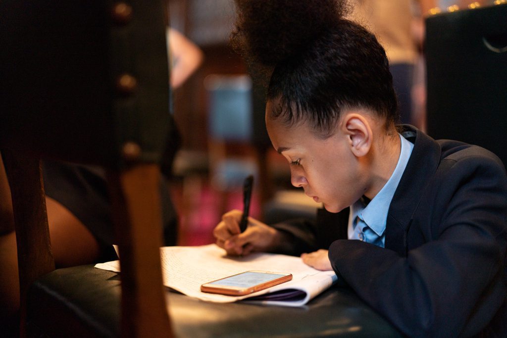 A young Black girl sits hunched over with paper and pen, concentrating on a speech she has written and is about to give at the Houses of Parliament