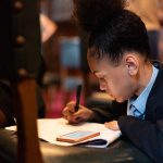 A young Black girl sits hunched over with paper and pen, concentrating on a speech she has written and is about to give at the Houses of Parliament