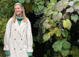 Erin Blackmore stands smiling outside on some grass, in front of a green, leafy bush. She has long, blonde hair that goes past her shoulders and is wearing a light green roll neck and beige trench coat with blue jeans and adidas trainers.