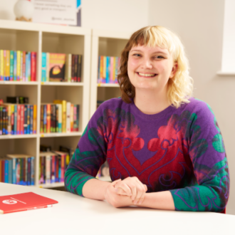 Rowan - a white woman with a fringe and half blond, half brown hair - site with her hands clasped in front of her. She wars a multicolour long sleeve top and smiles happily at the camera.