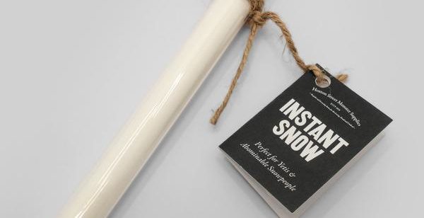 A long, clear plastic tube filled with white imitation snow, sealed with a brown cork. A black label hangs on hessian string and reads 'Instant Snow'.
