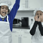Two students, dressed in chef aprons and hats, cheer loudly and throw their arms up in joy