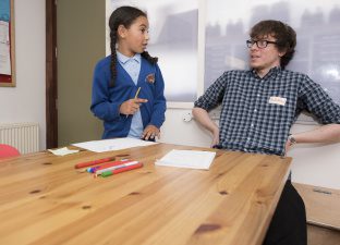 A young male writing mentor sits in a checked shirt looking surprised and happy with what a young writer - who is standing and explaining a story - is describing to him.