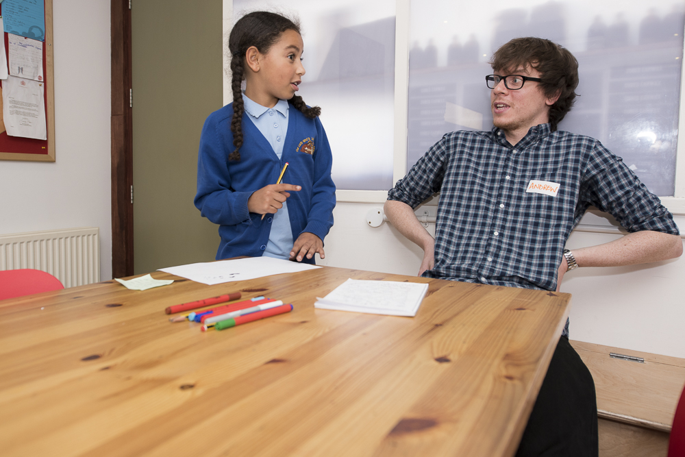 A young male writing mentor sits in a checked shirt looking surprised and happy with what a young writer - who is standing and explaining a story - is describing to him.