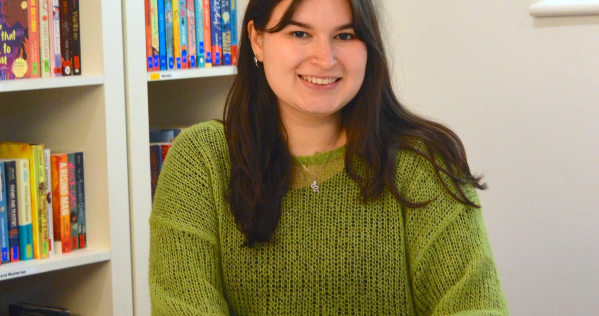 Tegan is a young white woman with long dark hair past her shoulders, she is sitting at a desk smiling with her hands clasped in front of her. She is wearing a green jumper with a small snake necklace.