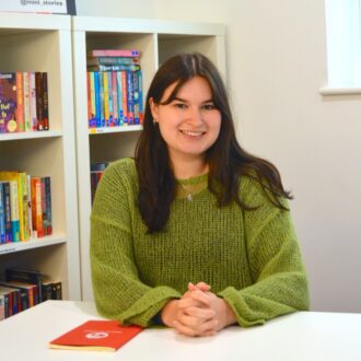 Tegan is a young white woman with long dark hair past her shoulders, she is sitting at a desk smiling with her hands clasped in front of her. She is wearing a green jumper with a small snake necklace.