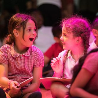 Two pupils look at each other excitedly. Each holds a pencil and notebook.