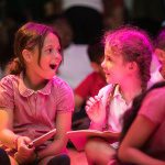 Two young girls, hair in plaits, wearing school uniform dresses, sit on the floor writing and excitedly exchange ideas