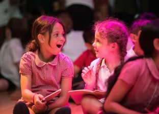 Two young girls, hair in plaits, wearing school uniform dresses, sit on the floor writing and excitedly exchange ideas