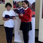 Two primary pupils smile as they read from their published anthology. in front of a Ministry Of Stories banner.