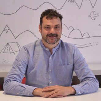 Justin wears a light blue shirt, has short dark hair and a short dark brown beard. He sits smiling with his hands clasped in front of him on a white desk.