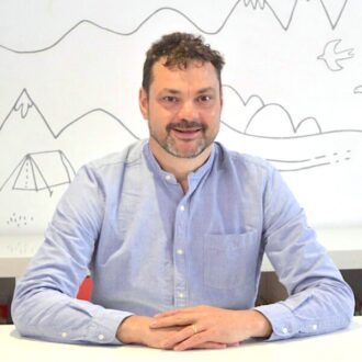 Justin is a white man wearing a light blue shirt, he has short dark hair and a short brown beard. He sits smiling with his hands folded in front of him on a white desk.