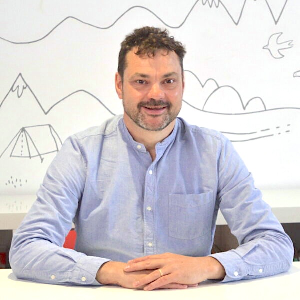 Justin is a white man wearing a light blue shirt, he has short dark hair and a short brown beard. He sits smiling with his hands folded in front of him on a white desk.