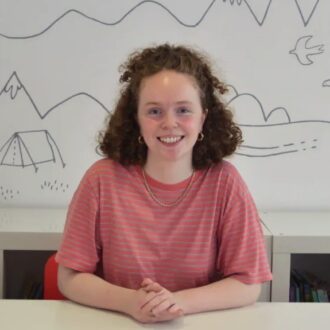 Phoebe Wagner has red hair in tight curls and is wearing a red stripy t-shirt with gold jewellery. She sits smiling behind a white desk hands clasped together.