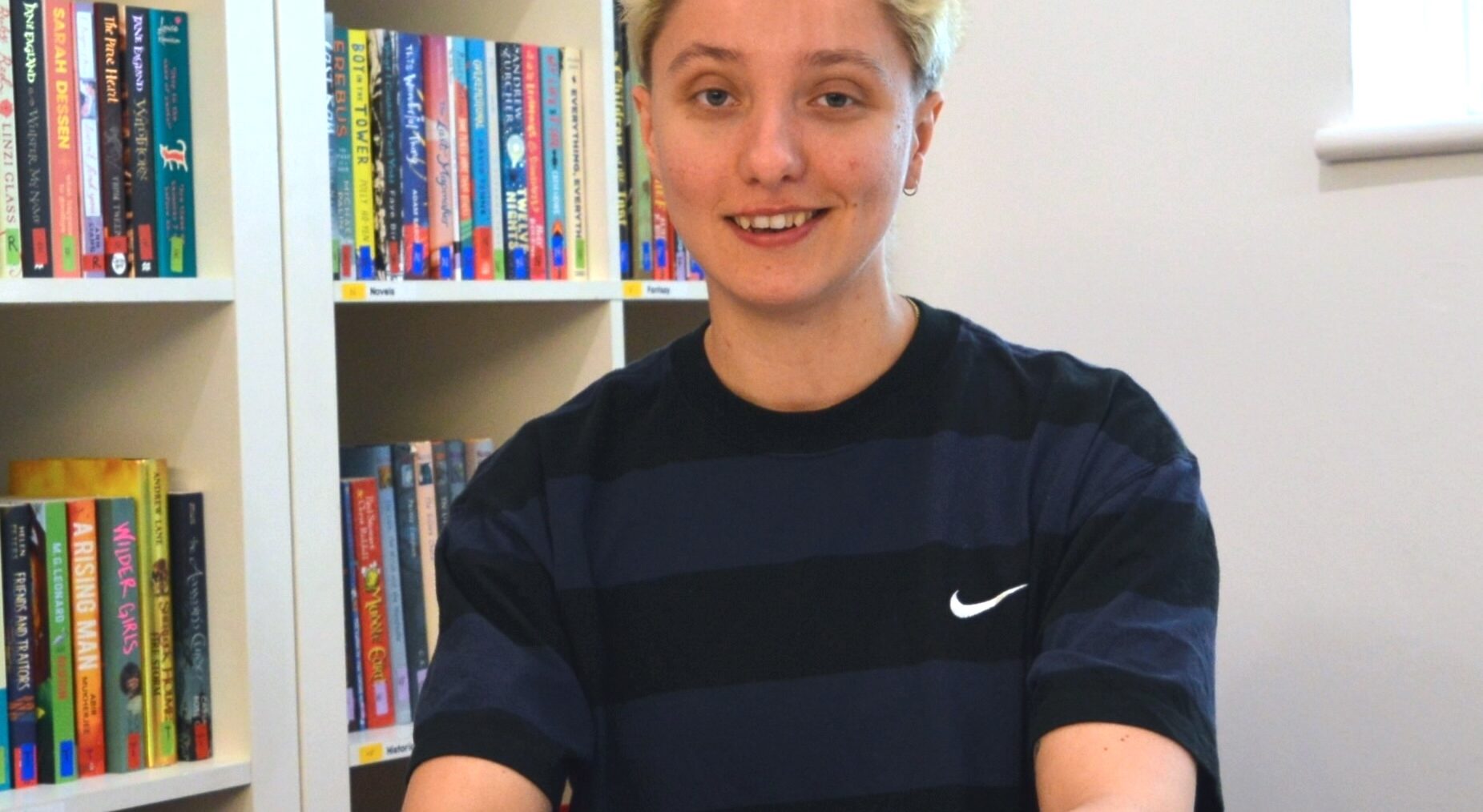Beattie is a young, white person with short spiky hair. They're sitting behind a desk wearing a blue and black striped t-shirt and smiling at the camera. Their hands are folded on the desk in front of them.
