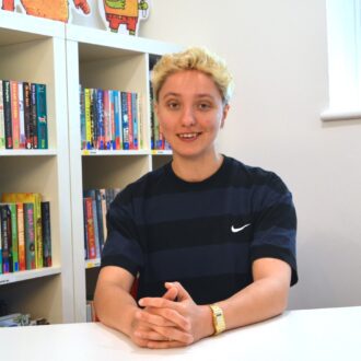 Beattie is a young, white person with short spiky hair. They're sitting behind a desk wearing a blue and black striped t-shirt and smiling at the camera. Their hands are folded on the desk in front of them.