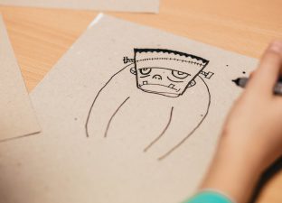 A childs hand rests, pen in hand, on an ink drawing of Frankenstein monster