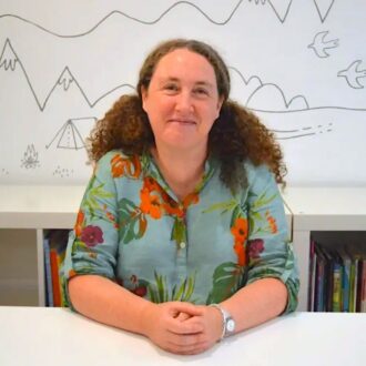 Becky is a white woman with long curly brown hair that spills over her shoulders. She is smiling at the camera with her hands clasped on a white desk. Becky is wearing a light blue shirt patterned with green leaves and blooming red and purple flowers.