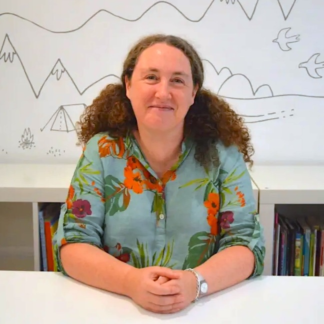 Becky is a white woman with long curly brown hair that spills over her shoulders. She is smiling at the camera with her hands clasped on a white desk. Becky is wearing a light blue shirt patterned with green leaves and blooming red and purple flowers.