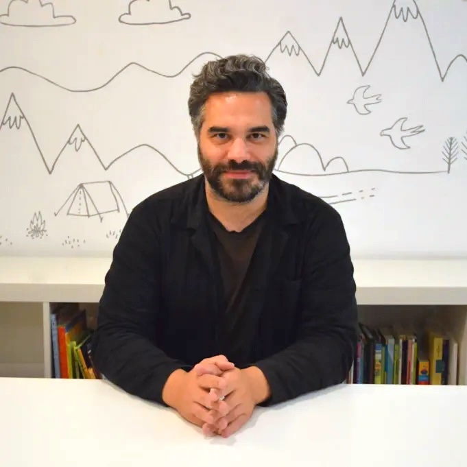 Michael sits with his hands folded on a white desk. He is a white man with short, styled black hair with flecks of grey above his ears. He has a short beard and is wearing a black jacket over a brown t-shirt.