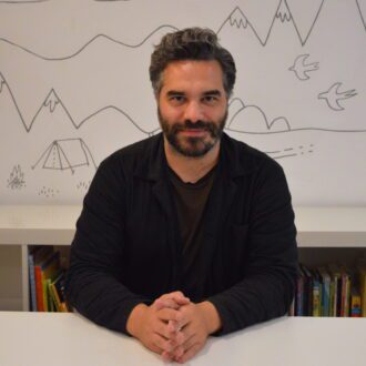 Michael Crowe sits with his hands folded on a white desk. He has short, styled black hair with flecks of grey above his ears. He has a short beard and is wearing a black jacket over a brown t-shirt.