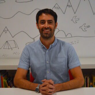 Michael sits smiling broadly with his hands intertwined on a white desk. He has short, dark brown hair and a short beard. He is wearing a light blue, short sleeved shirt.