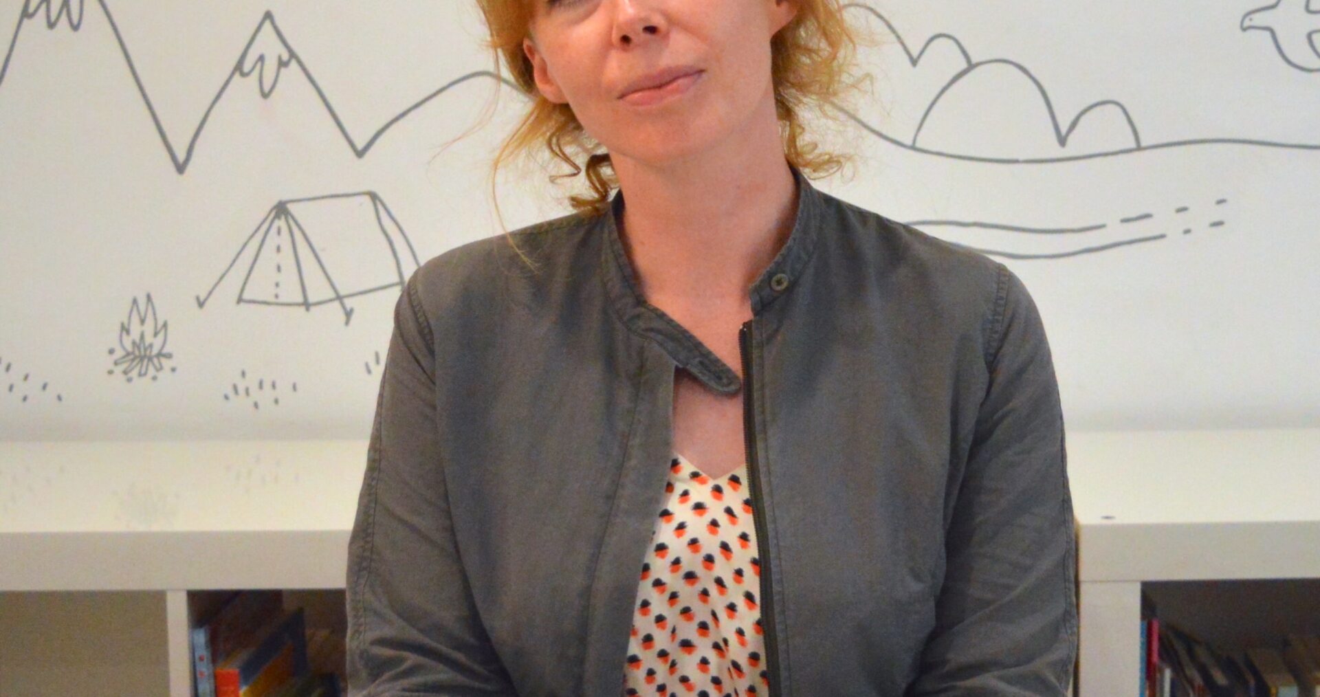 Michelle is a white woman with orange hair. She is sat in front of a book case and white wall with doodles of mountains, trees and birds on it. She is wearing a red and white polka dot dress and a grey jacket and is smiling at the camera with her hands folded on the desk in front of her.