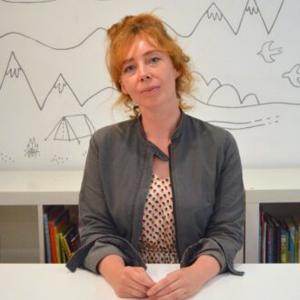 Michelle is a white woman with orange hair. She is sat in front of a book case and white wall with doodles of mountains, trees and birds on it. She is wearing a red and white polka dot dress and a grey jacket and is smiling at the camera with her hands folded on the desk in front of her.