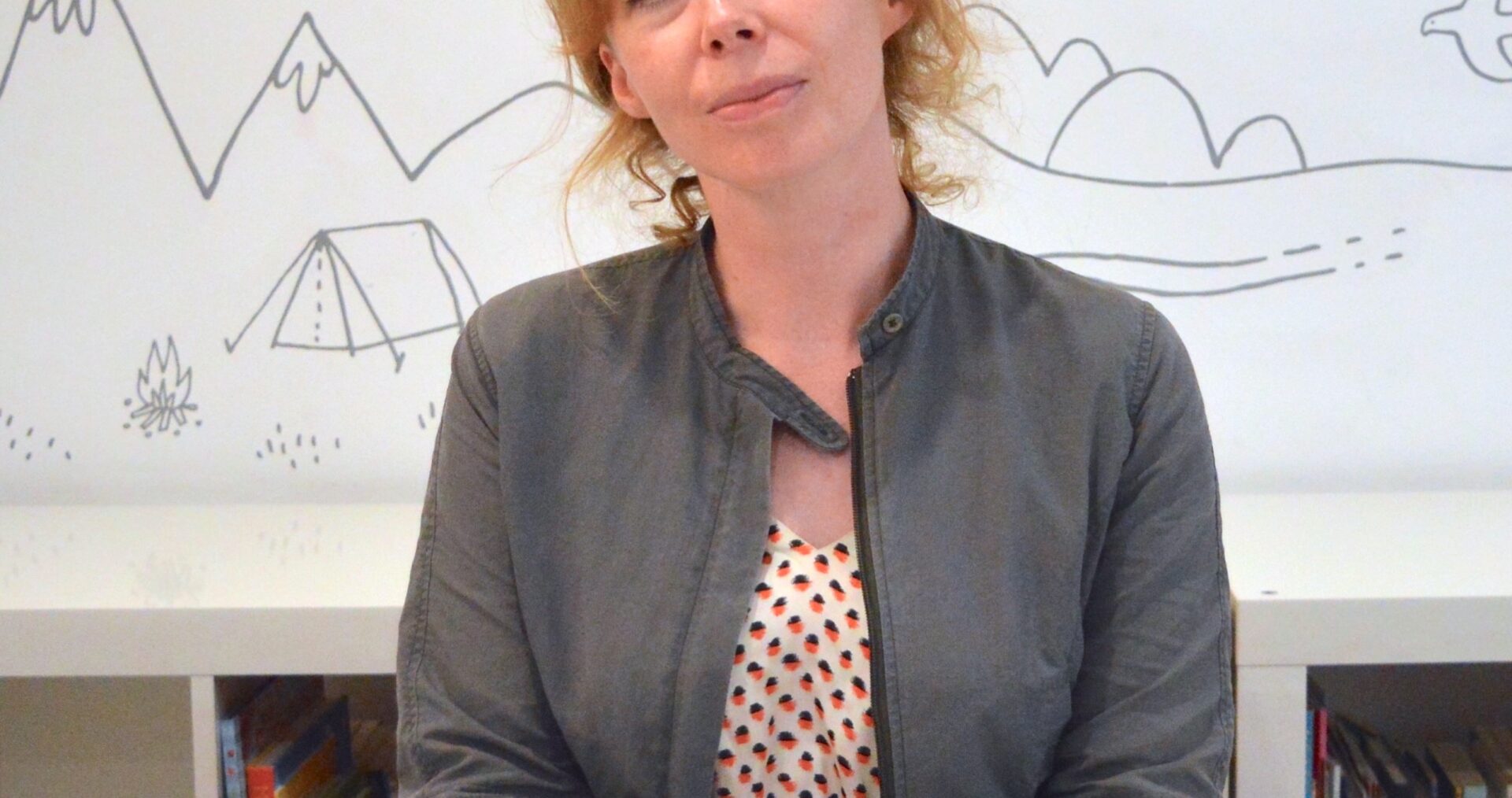 Michelle is a white woman with long, curly ginger hair. She is sat in front of a book case and white wall with doodles of mountains, trees and birds on it. She is wearing a red and white polka dot dress and a grey jacket and is smiling at the camera with her hands folded on the desk in front of her.