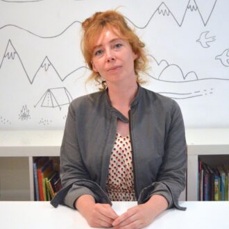 Michelle is a white woman with long, curly ginger hair. She is sat in front of a book case and white wall with doodles of mountains, trees and birds on it. She is wearing a red and white polka dot dress and a grey jacket and is smiling at the camera with her hands folded on the desk in front of her.