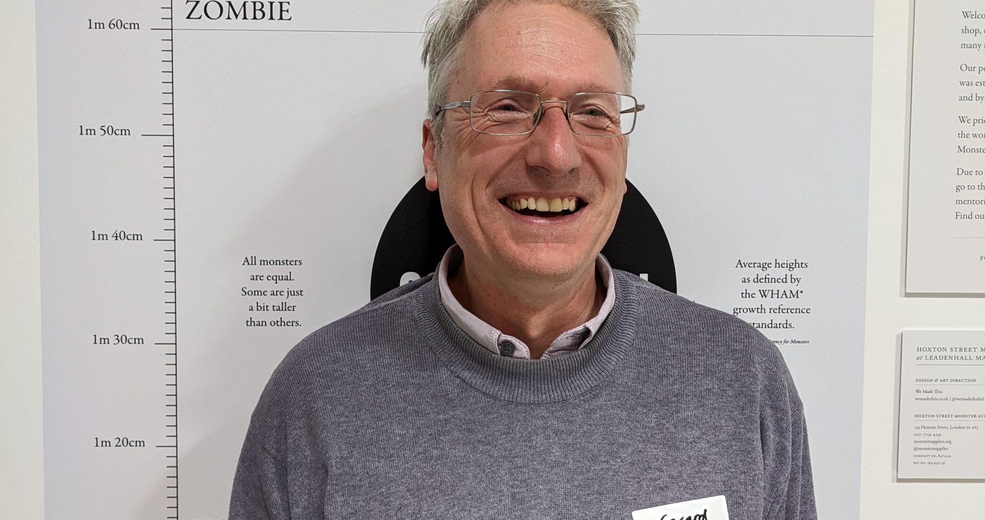 Gerard Darby stands smiling at the camera in front of a Hoxton Street Monster Supplies height chart. He wears a grey jumper and silver rimmed glasses.