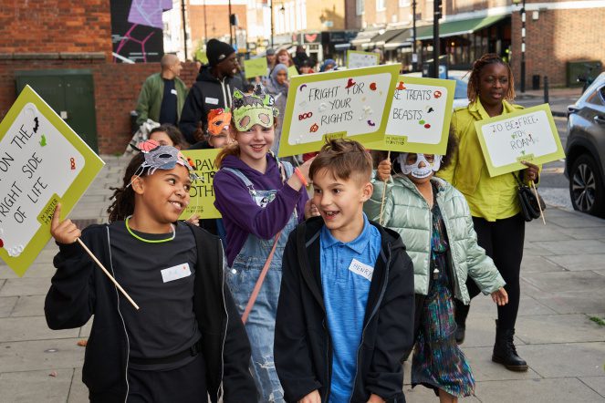 A group of young writers smile and laugh as they walk down Hoxton Street surrounded by placards and hand drawn monster masks