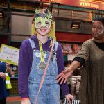 A young writer stands smiling against a red shop wall, wearing dungarees and a green, hand coloured monster mask and a lime green Monster Month medal.