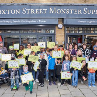 A large group of young people and adults stand in front of the grey Hoxton Street Monster Supplies shop waving lime green banners and placards with fun, monster puns written in black ink.