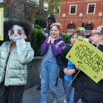 Three young writers walk smiling and laughing together wearing with a placard that reads 'one paw in front of the other'