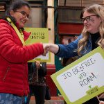 Two young writers, in a red and blue winter coat, smile and fist bump each other in from of the monster shop