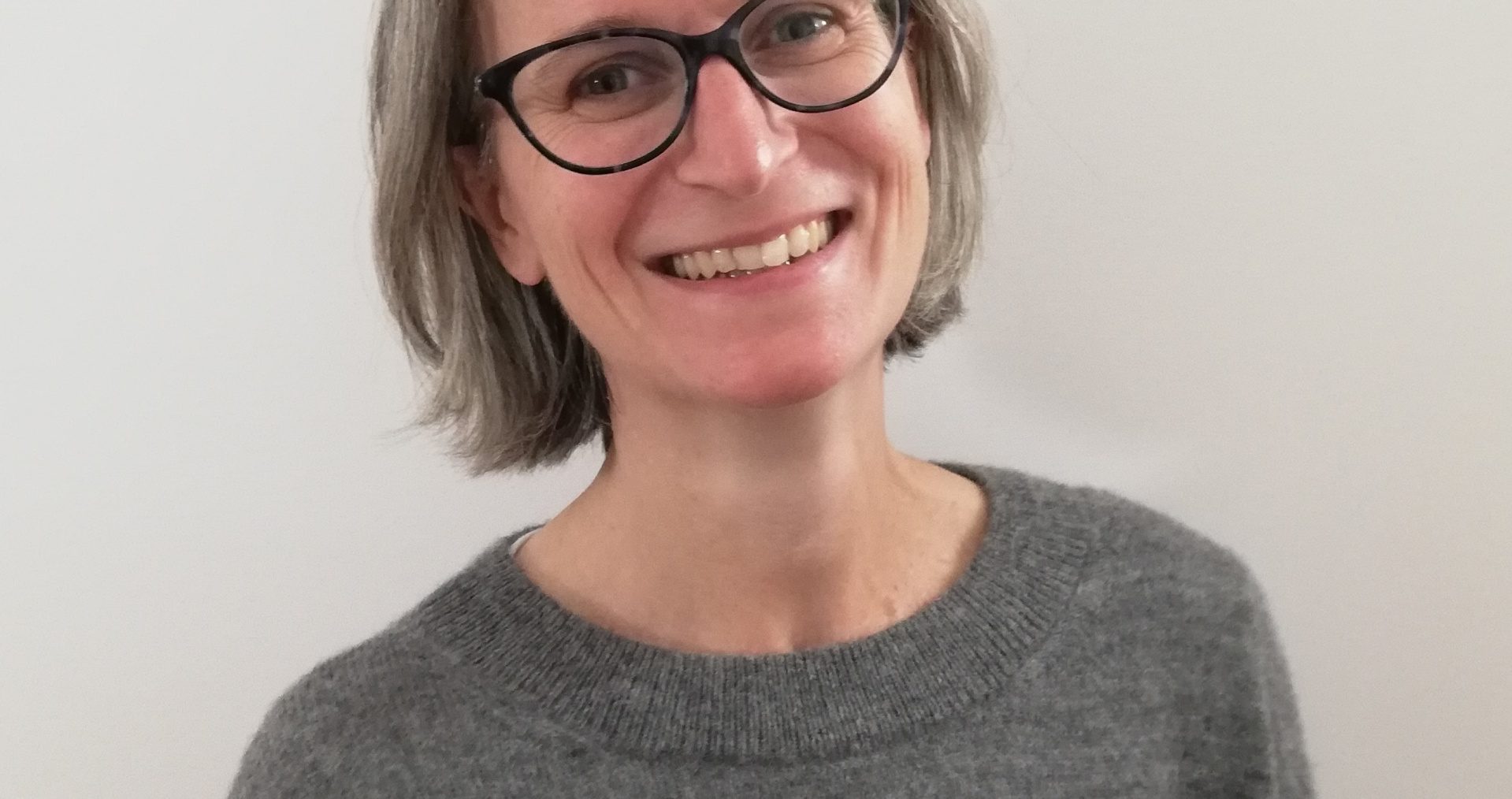 Nicole Gurvidi sits smiling at the camera. She wears a grey jumper and black rimmed glasses.