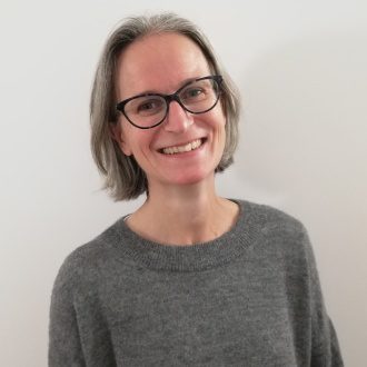 Nicole Gurvidi sits smiling at the camera. She wears a grey jumper and black rimmed glasses.