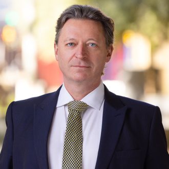 Philip Watkins sits facing the camera in a relaxed manner wearing A yellow tie, white shirt and blue suit.