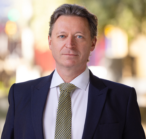 Philip Watkins sits facing the camera in a relaxed manner wearing A yellow tie, white shirt and blue suit.